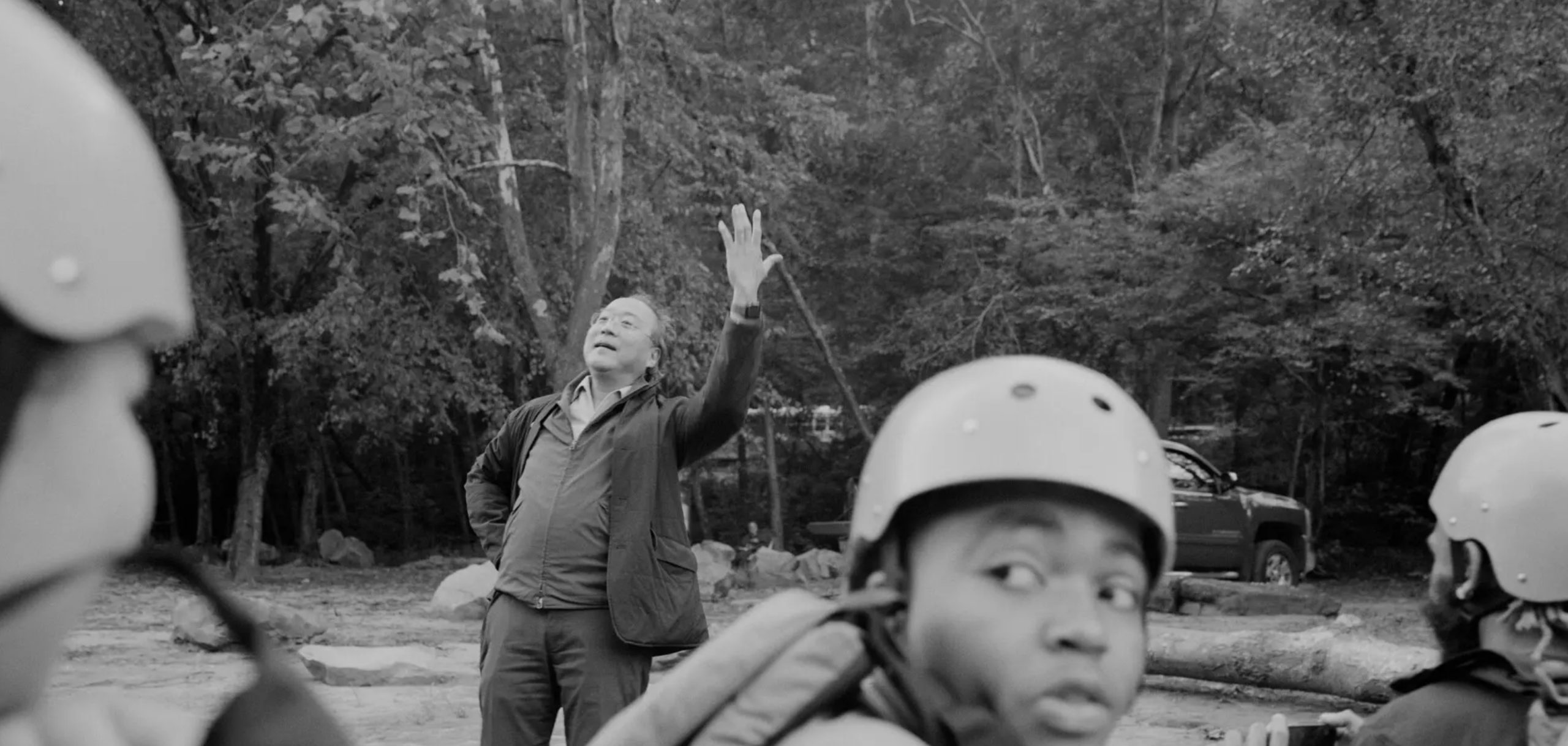 Black and white photograph of Yo-Yo Ma, casually dressed, standing in forested area with several boys wearing helmets for exercise on Ronnie Stangler good reads page regarding Yo-Yo Ma Finding His Way Back to Nature Through Music.